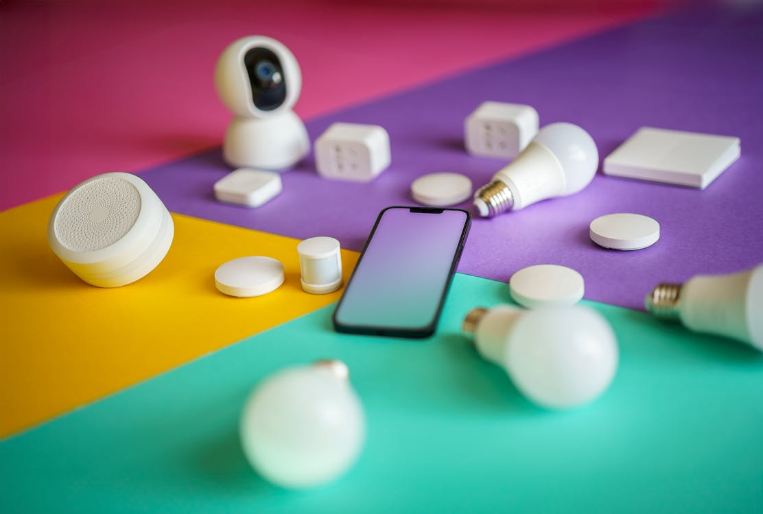 Smart home devices including bulbs, plugs, sensors, and a smartphone on a colorful background representing home automation.