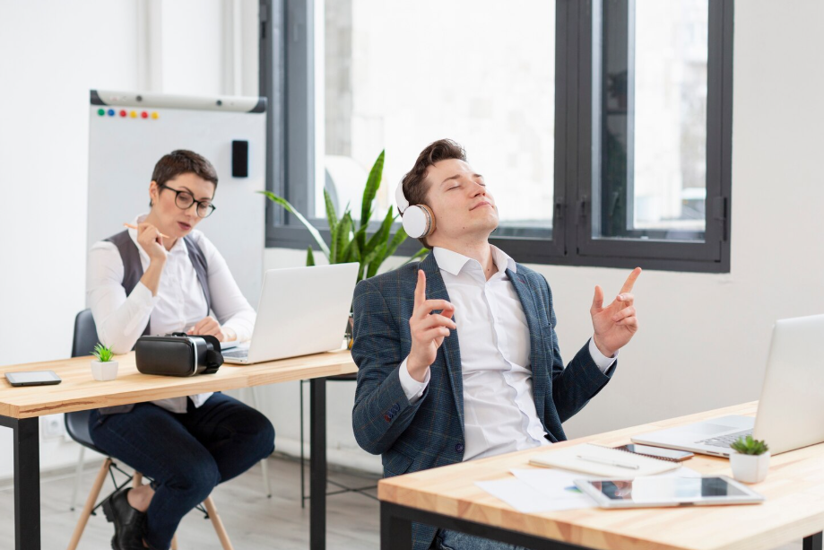Office worker enjoying music with headphones while colleague works on a laptop, reflecting balance between focus and relaxation at work.